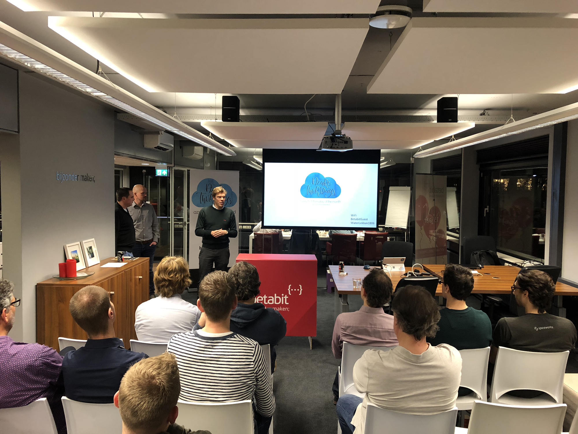 A host standing at the front of a small meetup room welcoming the audience, the title slide and the wifi password still on screen